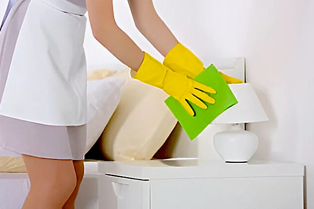 A lady wearing yellow gloves cleaning a white table lamp with a green cloth